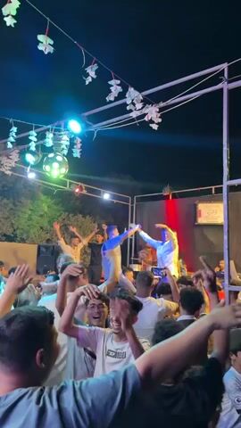 A nighttime outdoor event is depicted with a raised stage in the background. A crowd of people, predominantly young males, are gathered and dancing in front of the stage. The stage is illuminated by colorful lights, including green and purple spotlights, and a disco ball is visible. Several individuals are on the stage, appearing to perform or entertain the crowd, with one person wearing a white shirt and another a blue shirt. Decorative elements, possibly flowers, are strung across overhead structures. The overall atmosphere suggests a lively celebration or party.