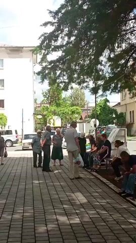 A group of people are gathered on a paved walkway in front of a building with a white facade and shops on the ground floor. Some individuals are standing and conversing, while others are seated on a bench. There are trees with green foliage providing shade. Cars are parked in the background. The time is daytime.