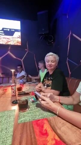 Four people are seated at a wooden table in a dimly lit room with blue lighting. On the wall behind them, a large screen displays an image of food. In the foreground, a woman with short gray hair and a black t-shirt smiles and holds a smartphone. To her left, a person wearing a light green tank top appears to be playing a game on a device. Further to the left, a man in a white t-shirt is waving. Another person, only partially visible, is seated to the right of the woman in the black t-shirt. Several drinks and cigarette packs are on the table. The atmosphere appears to be casual and social.