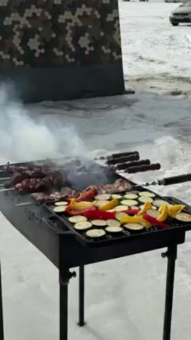 A low-angle shot shows a black charcoal grill in a snowy, outdoor setting. On the grill, skewers of meat are cooking alongside sliced vegetables, including zucchini, red bell peppers, and yellow bell peppers. Smoke rises from the grill, obscuring the meat somewhat. Behind the grill, there is a camouflage-patterned structure, likely a tent or shelter. In the background, several cars are visible in a parking area. The scene is bathed in bright daylight.