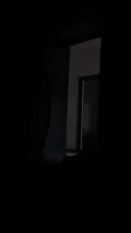 The video shows a view into a room from a dark space. A light fixture is partially visible in the room, which appears to be white. There is an open doorway into another room. A curtain hangs to the left. The scene is dimly lit and the exact details of the objects in the room are difficult to make out.