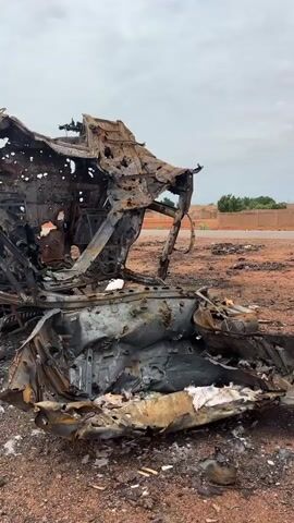 The video shows the aftermath of what appears to be a vehicle fire or explosion. A heavily damaged, burnt-out vehicle, possibly a truck or large SUV, is the central focus. The chassis and frame are severely warped and blackened, with metal twisted and melted. The body panels are mostly gone or heavily damaged, revealing the internal structure. Debris, including ash, melted material, and possibly shrapnel, is scattered around the vehicle on a dirt or gravel surface. In the background, a low, dusty landscape is visible with some sparse vegetation and a building or structures in the distance. The sky is overcast. There are no visible people in the immediate vicinity of the vehicle, though a few figures can be seen further in the background near the structures. The scene suggests destruction and abandonment.