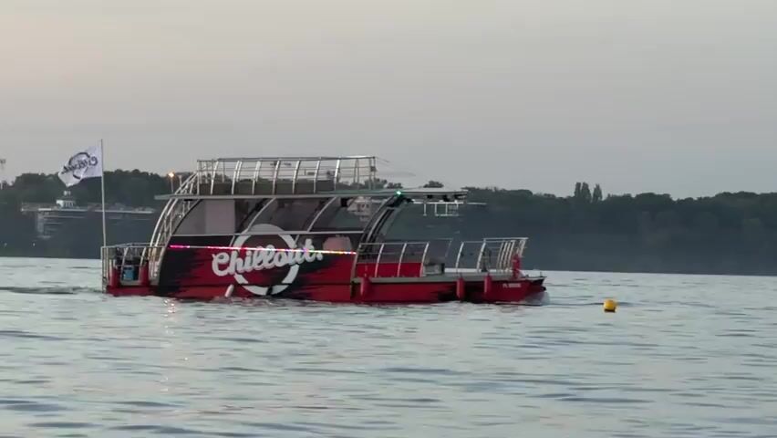 A red and black boat with "Chillout" written on the side is moving through the water. The boat has a roofed area with windows and an open deck area. There are several yellow and red buoys in the foreground. The sky is overcast.