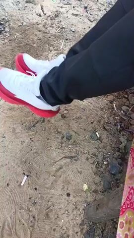 A close-up shot focuses on a person's lower legs and feet, wearing white sneakers with red soles and black pants. The person is sitting, and their feet are tapping rhythmically on a sandy surface littered with small stones, twigs, and cigarette butts. In the background, a glimpse of a floral-patterned fabric is visible, suggesting the presence of another person or an object. The lighting suggests an outdoor setting during the day.