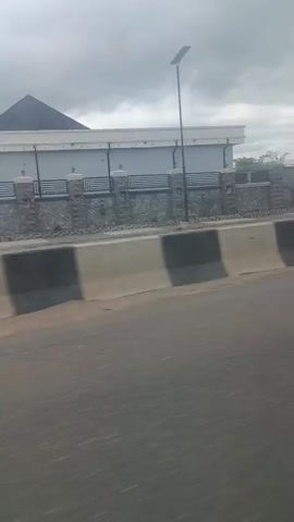 The video shows a view from a moving vehicle on a road. The road is paved and has a barrier made of concrete blocks painted in alternating black and beige sections. Beyond the barrier, there is a wall with a decorative pattern and a fence topped with barbed wire. A street light is visible on a pole in front of a modern-looking building with a dark pitched roof. Further down the road, trees and vegetation are visible, along with more of the decorative wall and fence. The sky is overcast. There are no people or animals visible in the frame.