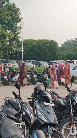 The video shows an outdoor parking lot, likely at a train station or bus station, given the presence of numerous motorcycles, scooters, cars, and people with luggage. Several individuals are visible walking and interacting with their belongings. In the foreground, a row of parked motorcycles and scooters is prominent. In the middle ground, several people are walking or standing with luggage, and some are engaged in conversation or seemingly waiting. There are also cars parked in the background, and trees and greenery beyond the parking area. The overall atmosphere appears to be one of travel and transit.