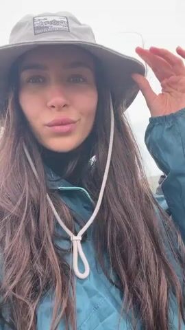The video shows a woman with long brown hair wearing a grey bucket hat with the words "Travel Never Ends" stitched on the front. She has on a blue jacket and is looking directly at the camera in a selfie style shot. Behind her stands a man wearing a black jacket and pants, a baseball cap, and red sunglasses. They are standing on a rocky outcrop with a view of the ocean and a grassy coastline. The sky is overcast and appears to be foggy. The man in the background gestures to the landscape.