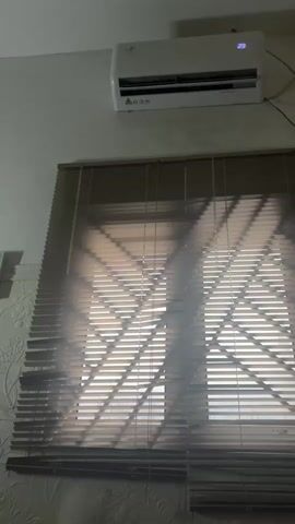 The video shows a split air conditioning unit mounted on a wall above a closed window with horizontal blinds. The air conditioning unit is white and has a digital display showing the temperature "29". The blinds are closed and appear to be a light brown color. Sunlight is visible through the blinds, casting geometric shadows on the window. The wall has a textured pattern. The video is still and appears to be a short loop.