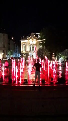 A person stands in front of a fountain with red lights at night. The fountain shoots water streams that are illuminated by red lights, creating a vibrant display. In the background, a building with lights on is visible, along with some trees and other structures. Other people can be seen in the distance. The overall scene depicts a lively urban environment at night.