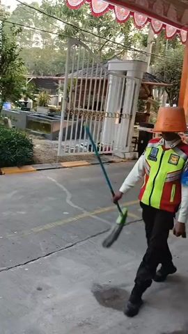 A person wearing a security uniform with a reflective vest over a tan shirt and black pants walks down a paved path. They have an orange bucket on their head, and a mop is slung over their shoulder. The person is walking away from the camera, toward the right side of the frame. The environment appears to be outdoors, possibly in a residential or commercial area, with trees, bushes, and a fence visible in the background. The lighting suggests it is daytime.