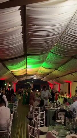 This video captures a lively reception held under a draped tent. The ceiling is adorned with fabric that creates a wavy effect, illuminated by strings of fairy lights and several ornate chandeliers. The lighting shifts through a spectrum of colors, from vibrant green to warm oranges and reds, creating a festive and dynamic atmosphere.

The scene is filled with people mingling and dancing. In the foreground, tables are set for a meal, suggesting a celebratory event like a wedding or anniversary. Guests are dressed in semi-formal attire, with women in dresses and men in shirts and trousers. Some individuals are actively dancing on a wooden floor, while others are seated at tables or socializing.

The overall mood appears joyful and celebratory. The combination of the colorful lighting, the elegant decor, and the presence of guests enjoying themselves points to a special occasion. While the specific time of day isn't explicit, the indoor setting with artificial lighting suggests it could be evening or late afternoon, depending on the venue's setup. The location is identified as Baliovtsi, Bulgaria. There is no visible text in the video.