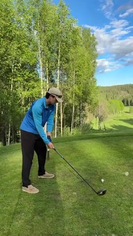 Here's a description of the video:

A man wearing a blue long-sleeve shirt, black pants, a tan cap, and tan shoes is seen on a golf course. He's teeing off, swinging his golf club at a golf ball.  After hitting the ball, he watches its trajectory. The background features a bright blue sky with scattered clouds, tall birch trees, a green fairway, and a distant, tree-covered hill.