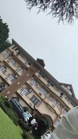 The video begins with a wide shot of a multi-story building with a beige facade and brown trim. The building has numerous windows, many with arched tops, and balconies. A sign on the building reads "IMPERIAL" upside down. To the left of the building is a well-manicured green lawn with bushes and trees. Several cars are parked in front of the building, including a black SUV, a white van, and a white SUV. A person's legs and torso wearing light-colored clothing are visible in the foreground, partially obscuring the view of the building and cars. The person is wearing a watch on their left wrist. The camera angle is tilted, making the scene appear sideways. The sky is overcast. The video appears to be a selfie-style recording, with the camera likely held by the person in the foreground.
