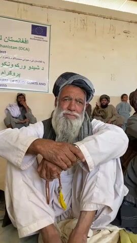A man with a long white beard and a turban is sitting on the floor, facing the camera. He is wearing a white shirt and pants. He is holding prayer beads in his hands. Behind him, several other men are sitting on the floor, and a banner with writing in a foreign language is hanging on the wall. The environment appears to be an indoor space with a concrete floor and a light-colored wall. The overall scene suggests a community gathering or meeting.