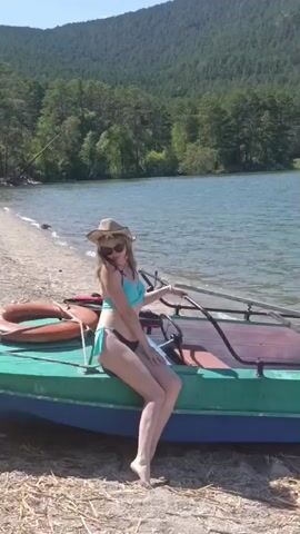 A woman in a bikini and a straw hat sits on the edge of a green and blue boat on a sandy beach. She is looking at the camera with a smile. Behind her is a large body of water, and in the distance, there is a wooded mountain. The boat has oars and a Yamaha motor attached to the back. The overall scene is one of a peaceful summer day by the lake.