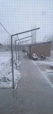The scene depicts a snowy, possibly rural landscape, viewed from a slightly elevated position, likely a porch or step. A metal structure with a series of horizontal bars extends into the foreground, possibly a trellis or support for plants. The ground is covered in snow, and there is a clear path made of concrete or stone leading into the distance. On the right side of the path, there's a small brick or stone building with a dark-colored roof. A wooden structure resembling a bench or stacked wood sits near this building. In the background, a blurred outline of another building is visible. The sky is overcast and bright, indicating a wintery day with snowfall.