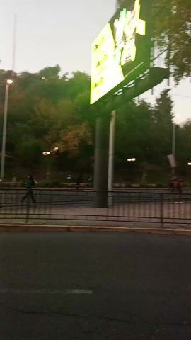 The video depicts a gathering of people in what appears to be a park or public square. A black metal fence separates the gathering from a road. Streetlights illuminate the park, and buildings are visible in the background. The people are moving around, and there is some activity. The overall tone of the scene suggests a public event or demonstration. Siren sounds can be heard.