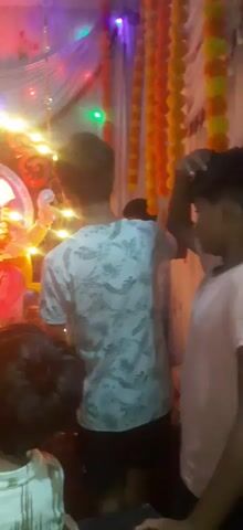A group of children and young people are gathered around a decorated altar for the Hindu deity Ganesha. The idol of Ganesha is adorned with flowers and sits in the center of the scene, illuminated by numerous strings of bright, warm-toned lights and some colored bulbs. Yellow and orange marigold garlands hang in the background, adding to the festive atmosphere. The individuals are facing the idol, with some looking directly at it and others looking towards the camera. One young person in the foreground is looking at the camera and appears to be speaking. The scene is dimly lit, with the lights from the altar being the primary source of illumination.