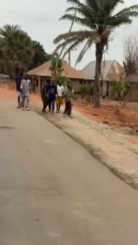 The video depicts a group of people walking along a paved road in what appears to be a rural or semi-rural setting. There are seven individuals visible: two older men, one wearing a light-colored, long-sleeved shirt and dark shorts, and the other in a dark shirt and shorts, are walking alongside a group of five younger individuals, possibly children. The children are wearing a variety of clothing including a blue shirt, white shirt, and green shirt. The background includes houses with corrugated metal roofs, trees, and an unpaved area alongside the road. The lighting suggests it is daytime, and the overall atmosphere seems calm.