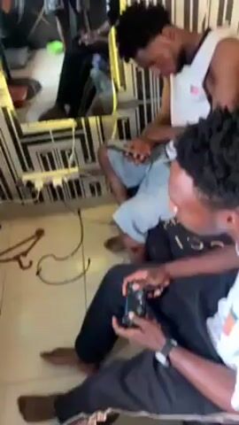 The video shows three young men indoors. One man sits with a game controller in his hands, seemingly engaged in playing a game. Another man is seated nearby, focusing on a mobile phone. The third man is partially visible, with his face down and appears to be deep in thought or emotional. A decorative mirror on the wall reflects the scene, capturing one of the men standing, posing for a picture with his phone. The room has a patterned wallpaper and the floor is visible with wires and objects strewn around. The lighting is relatively dim, but the mirror is lit up. There is music playing in the background.