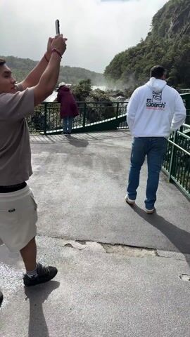 The video shows a scenic view of a river from a bridge. Initially, a man with a phone is visible, taking a photo of the landscape. He wears a brown shirt, light-colored shorts, and red wristbands. Another man in a white hoodie and jeans walks past him. The camera pans to reveal a green bridge with multiple people on it, surrounded by lush greenery. Following this, the camera shows a rapid, turquoise river flowing amidst the forested landscape. The environment appears mountainous with dense vegetation.