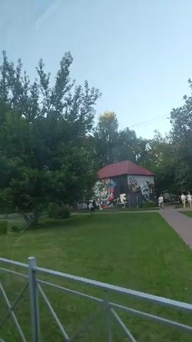 This video captures a moving view of a park-like area, likely in Ramenskoye, Russia. The time of day appears to be late afternoon or early evening, with soft, diffused sunlight and a bright, overcast sky. The scene is dominated by lush green grass and a variety of trees, some in full leaf, suggesting a warm season like summer or late spring.

In the foreground, a silver metal railing with a crisscross pattern is visible, indicating the perspective is from behind this barrier, possibly from a moving vehicle or a walkway.

Several objects stand out. A prominent traffic sign, a blue square with a white triangle inside and a black shape representing a speed bump, warns drivers of an upcoming obstruction. Beside it, a circular speed limit sign displays "20" in red text on a white background, indicating a maximum speed of 20 kilometers per hour. Further in the background, a large billboard stands on two dark pillars. Part of the text on the billboard is visible and appears to be in Cyrillic script. One section clearly reads "МЕСТО СВОБОДНО" which translates to "PLACE IS FREE". This suggests the billboard might be advertising available space.

A building with a red roof is visible behind the trees, adorned with colorful murals or graffiti depicting various images, including what appears to be a portrait of a man and some abstract designs. Some people are seen walking in the distance, adding a sense of gentle activity to the scene. The overall atmosphere is peaceful and somewhat serene, with a hint of urban life subtly integrated into the natural setting.