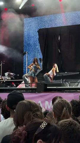 The video shows a concert performance from behind a crowd of spectators. On a brightly lit stage, three young women are performing. The central performer, wearing a denim skirt and top, holds a microphone and sings. Two other women, dressed similarly in crop tops and denim shorts, are positioned to her left and right, performing synchronized dance moves. The stage backdrop features a large screen displaying a textured blue pattern, and black curtains hang to the right. Stage lighting includes bright white spotlights and red beams that cast a colorful glow on the performers and the smoke-filled air. The crowd in the foreground is mostly out of focus, but their hairstyles and the tops of their heads are visible, indicating they are looking towards the stage.