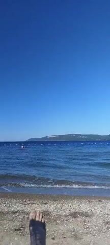 A person's legs and feet are visible in the foreground of the video, positioned on a beach with a towel underneath. The person is lying down, facing the ocean. The ocean is calm with gentle waves lapping at the shore. In the distance, there is a coastline with a hill or mountain. The sky is clear and blue, indicating a sunny day. There are some buoys or markers in the water, and a small boat or ship can be seen on the horizon. The video pans slightly to the right.