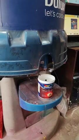 A paint-mixing machine, labeled "Dulux let's colour," is dispensing a liquid into a white can of Dulux Velvet Touch paint. The can sits on a small, round, blue-topped platform. The machine itself is blue and gray, showing signs of wear and tear. Behind the machine, a computer monitor displays an image, possibly related to the Dulux paint products. The surrounding area shows a cluttered environment with various objects and materials.