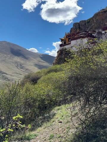 The camera pans slowly up the side of a mountain to reveal a monastery built into the cliff face. The monastery is white with red trim and a golden roof. The surrounding landscape is mountainous and rugged, with sparse vegetation. A blue sky with fluffy white clouds provides the backdrop. The shot emphasizes the monastery's isolation and integration with the natural environment. No people are visible.