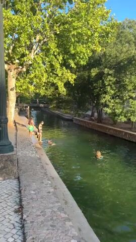 A short video shows a clear, green canal with people swimming and sitting on the edge. On the left bank, a person in a green and pink bikini stands on a stone pathway next to the canal. Further down the canal, another person is in the water, partially submerged. On the right bank, a person with blonde hair is swimming. Trees with green foliage line both sides of the canal, casting shadows on the water. Sunlight illuminates the scene, creating a bright and warm atmosphere.