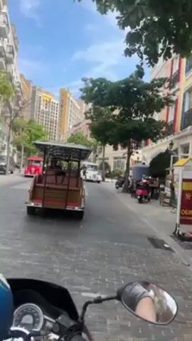 The video shows a first-person perspective of someone riding a scooter on a cobblestone street in a city. There are buildings on both sides of the street. A brown and a white tourist buggy are parked on the side of the road. There are also parked motorcycles on the right side of the road. The sky is blue with some clouds.