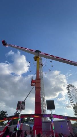 This video captures a vibrant amusement park scene on a bright, sunny day with a clear blue sky and scattered white clouds. The main subject is a large, tall amusement park ride, likely a pendulum or booster type, with a long arm that swings people high into the air. At each end of this arm, multiple people are seated, experiencing the thrill of the ride as it rotates and elevates them.

Adjacent to this large ride, a smaller swing carousel is also in operation, spinning people in individual swings. In the background, partially visible on the right, is a large Ferris wheel, further confirming the amusement park setting. Below the main swinging ride, two large screens are visible. One clearly displays the text "EXTREME" and parts of "BOOSTER". Another screen below appears to say "WELCOME ON BOARD". People are seen standing near the base of the rides, either waiting to participate or observing the attractions. The rides are illuminated with various colored lights, adding to the festive atmosphere. The overall impression is one of excitement and entertainment at a fairground, possibly located in Charenton-le-Pont, France.
