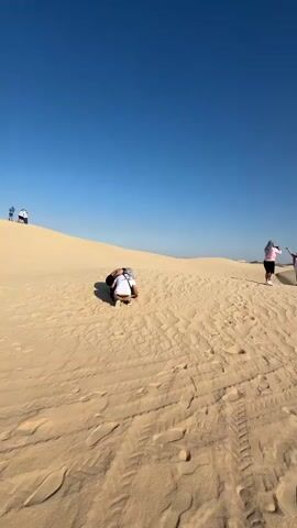 The video shows a group of people in a desert landscape under a clear blue sky. The environment consists of rolling sand dunes with visible footprints and tire tracks. Initially, three people are visible: one is crouched down, while the other two stand together, one raising their arms in the air. As the camera pans, a larger group of people comes into view, standing near several parked ATVs, some red and some black. Two people are dressed in traditional robes. The sun is shining brightly, and the scene appears to be in a remote, arid location.
