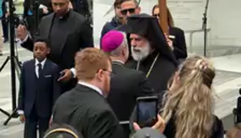 An outdoor scene captures a formal gathering, likely a religious event in New York, United States. The focal point is a man with a long white beard, dressed in black Orthodox Christian clerical robes and a black klobuk (headwear), holding a staff or cross. He is engaged in conversation with another man in a black suit, white shirt, and a distinct magenta skullcap (a zucchetto, typically worn by Catholic bishops). The bearded man appears animated, gesturing with his right hand while speaking, and the man in the magenta skullcap listens intently.

Around these central figures, a crowd is visible. To the left, a young boy in a dark suit stands beside a man in a black shirt with a clerical collar, and another man in a suit. Several other individuals, including what appear to be security personnel and members of the public, are present. To the right, a woman with long blonde hair holds a smartphone, seemingly recording the interaction. The background suggests a paved outdoor area, possibly a plaza or courtyard adjacent to a building. The lighting indicates it is daytime, and the weather appears clear. A religious chant is audible in the background, adding to the spiritual atmosphere of the event. No specific text is visible in the frame.