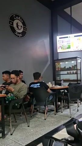 The video shows the interior of a coffee shop called "Zahir Kopi." Several people are seated at tables, some are looking at their phones, and others are facing away from the camera. A large television screen is mounted on the wall, displaying what appears to be a game or sports broadcast. There is a display case with various items, possibly food or merchandise, and a counter area with bottles and jars. A ceiling fan is visible. The overall atmosphere appears casual and relaxed.