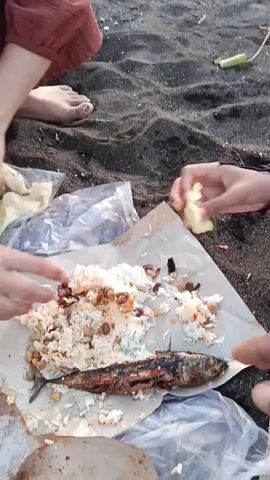 The video shows a close-up view of two people's hands and feet on a dark sandy beach. In the foreground, a plate of food is spread out on brown paper, consisting of rice, nuts, and a grilled fish. One person's hands are seen reaching for food. In the background, the ocean and a pier are visible, with a boat anchored in the water. The sky is a soft orange and pink, indicating either sunrise or sunset.
