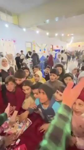 The video shows a crowd of excited children surrounding a person who appears to be distributing items, possibly gifts or treats. The children are of varying ages and ethnicities, and they are reaching out towards the distributor with enthusiasm. The scene takes place in a large, well-lit indoor space that may be a community center or event hall, decorated with balloons and streamers, suggesting a festive occasion. There are other people in the background, including adults, some of whom are wearing headscarves. The overall atmosphere appears to be lively and joyful.