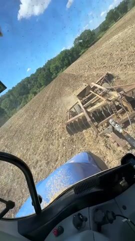 The video shows a view from inside a blue tractor cab as it plows a field. A disc harrow, attached to the tractor, is in operation, churning up the soil. The field appears to be dry and dusty, with some vegetation visible on the edges. The sky is blue with some clouds, and trees are visible in the background. The camera angle is high and looking down at the field. The action is continuous, with the tractor moving forward and the disc harrow working the soil. There are no people visible outside the cab. The lighting suggests it is daytime, likely mid-morning or afternoon. The overall scene depicts agricultural work in progress.