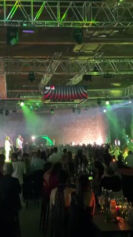 The video shows a dimly lit indoor event space with a large crowd of people seated at tables. Green spotlights illuminate a stage at the far end of the room where two people are performing. The stage is backed by a stone wall and a large screen displaying a red and black logo. The ceiling is adorned with exposed metal trusses, lights, and speakers. The audience members are facing the stage, some are standing and some are seated. The overall atmosphere appears to be lively and celebratory.