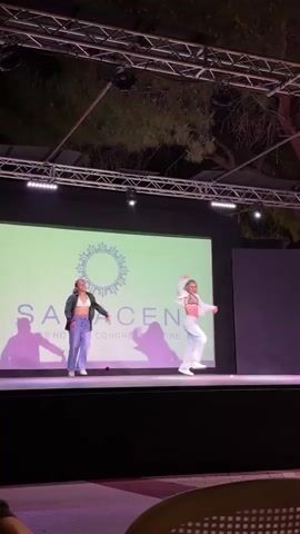Two young women with blonde hair are dancing on a stage. They are both wearing loose-fitting pants and crop tops, with one also wearing an open jacket. The woman on the left is wearing a dark jacket and grey pants, while the woman on the right is wearing a white outfit. They are performing in front of a large screen that displays a logo with the word "SARACEN" and text below it, along with stage lighting and a metal truss structure above. The background appears to be outdoors at night, with trees visible. The overall atmosphere suggests a performance or event.