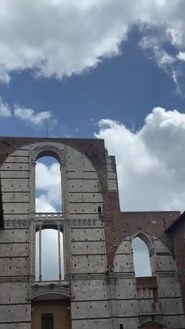 The video shows an architectural ruin against a blue and white cloudy sky. The ruin is made of stone and brick and has large archways. The top of the structure is incomplete. The architecture has dark accents across the facade.