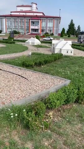 The video shows a miniature park with several small-scale buildings and landscaping. In the foreground, there are tiny white buildings with white roofs, surrounded by gravel pathways and green grass. In the background, a larger, more modern building with a red roof is visible. Trees and bushes are scattered throughout the park. The sky is overcast. The camera pans from left to right, showcasing the different models and the surrounding environment. The overall impression is that of a well-maintained miniature village or town.