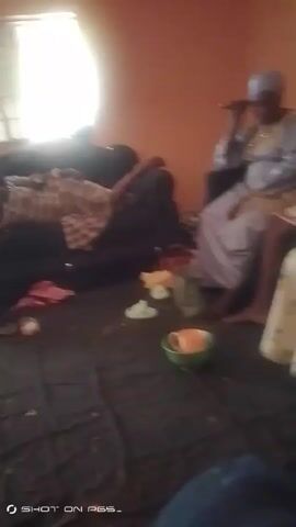 A woman in a light-colored dress and head covering sits on a gray couch, holding an object to her face. A person lies on the couch next to her. A table covered with a floral cloth is on the right. The floor is covered with a dark mat. A green bowl and other items are scattered on the floor. The room has light orange walls. The quality of the video is blurry.