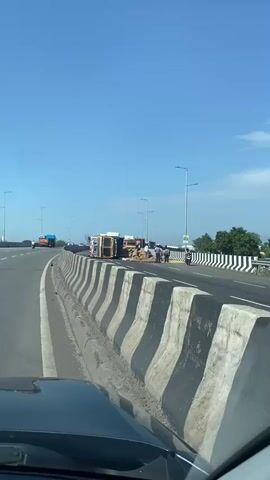 A vehicle is overturned on a highway, spilling its cargo of sacks. Several people are standing around the scene. The sky is clear and blue, and there are other vehicles in the background.