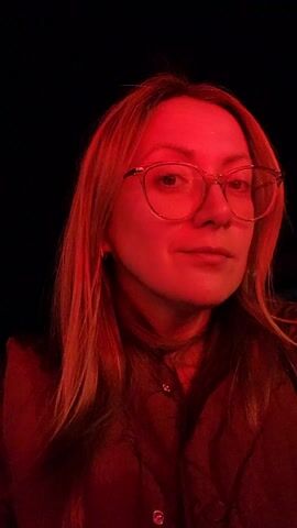 A woman with fair skin and reddish-blonde hair is centered in a close-up shot against a black background, illuminated by red light. She is wearing clear-framed glasses. She is wearing a button-down shirt and a jacket. She looks directly at the camera. Her facial expression transitions from neutral to a slight smile. The background is completely dark. The video is taken from a low angle, looking up at her.