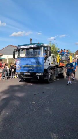 The video shows a parade float consisting of a white flatbed truck decorated with banners and plants. On the front of the truck, a blue banner displays German text, including "KOL-MEISTER UND Aufsteiger LK 2026" and "Euer Hip-Hop wird wahr: WIR sind wir". Several young men, likely a sports team, are standing and sitting on the back of the truck, wearing blue and white jerseys. Some are holding trophies or celebratory items. A man in a blue t-shirt and shorts is walking alongside the truck, looking at his phone. A crowd of spectators is visible on the left side of the road, standing behind a metal barrier. The scene appears to be a celebration, possibly a victory parade. The environment is outdoors with a blue sky and some clouds. The truck is on an asphalt road.