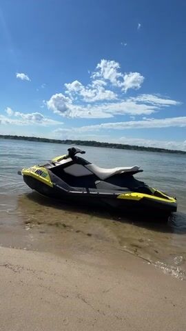 A jet ski is parked on a sandy beach with its rear in the water. The jet ski is black, yellow, and white. The sky is blue with white clouds. The water is calm. The overall scene is peaceful and serene.