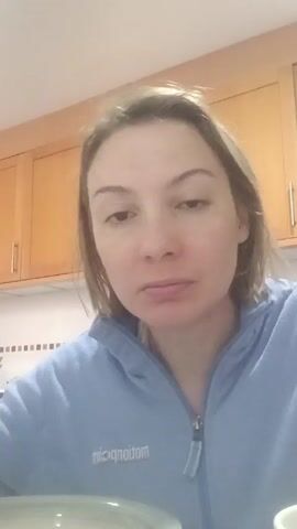 A woman is in a kitchen, wearing a blue zip-up hoodie. She is looking directly at the camera and eating from a spoon. The kitchen has light brown cabinets and a white tiled backsplash with tan and brown accents.