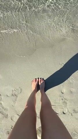 The video begins with a close-up, overhead shot of a person's bare feet standing on a sandy beach, with the edge of clear, shallow water lapping at the sand. Footprints are visible in the sand around the feet. The camera then pans up and slightly to the right, revealing the tranquil turquoise water with sunlight glinting off its surface. Several rocks, some partially submerged and covered in seaweed, are scattered in the water. In the distance, a coastline with hills and vegetation is visible under a clear blue sky. A few boats are anchored or moving slowly in the distance. The scene suggests a peaceful, sunny day at the beach.